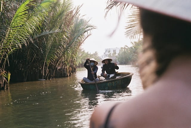 Deux femmes dans un bateau panier rond sur un canal au Vietnam, entourées de palmiers d'eau, observées par un passager à l'avant-plan. Une scène paisible évoquant les excursions aquatiques comme au lac de Ba Be.